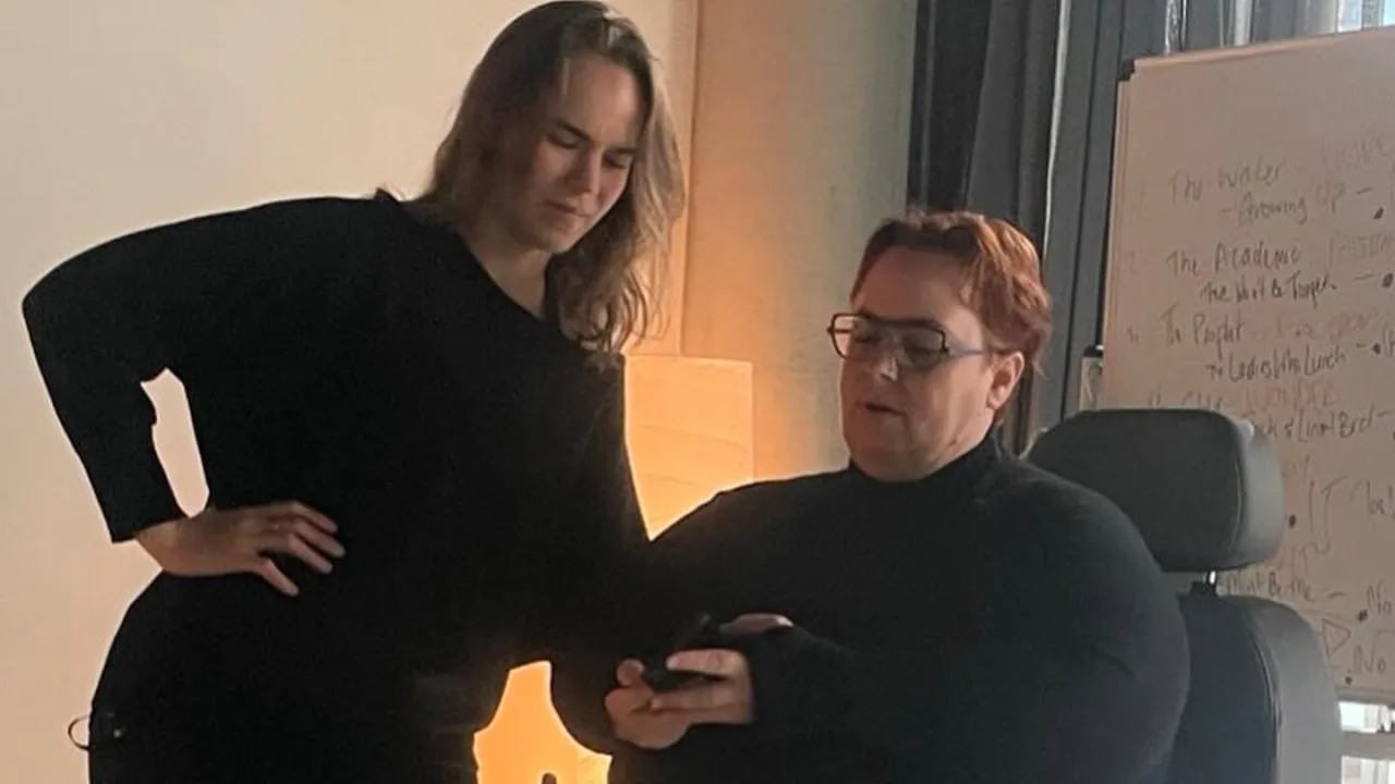 A white-skinned human with long brown hair, looks down at a device in the hands of a white-skinned human with short red hair, wearing purple glasses and sitting in a power wheelchair. They are both wearing theatre blacks. In the background is a large whiteboard with colourful writing on it