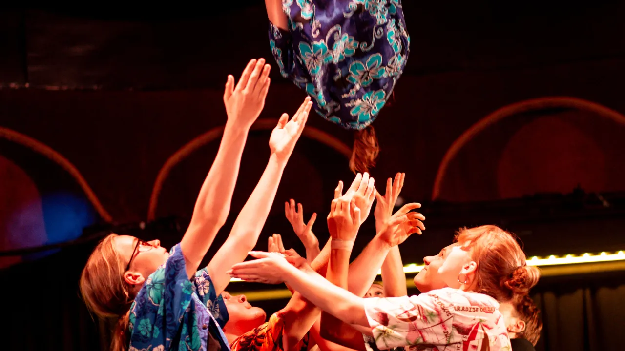 Six young circus performers in costumes of Hawaiian shirts stand closely together, arms reaching upwards, ready to catch their seventh member who is pictured upside down and falling towards them after flipping through the air. They are on standing on the dimly-lit stage of a Spiegeltent.