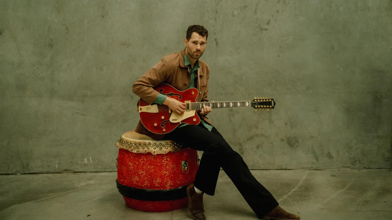 A contemplative musician sits on a weathered red ceremonial drum, holding a vintage red Gretsch guitar. Dressed in a brown jacket, green shirt, and dark trousers, he gazes toward the camera with quiet intensity. Against a raw concrete wall, the scene feels timeless and understated.
