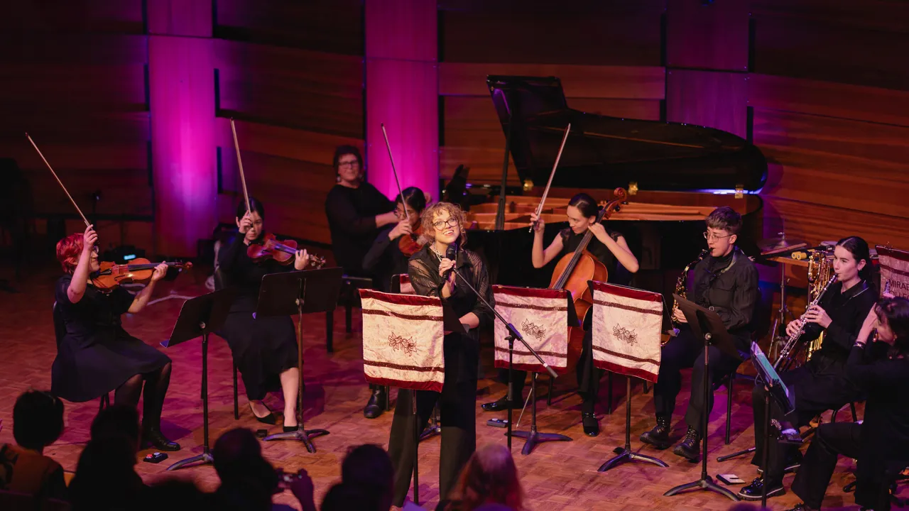 Mirabilis Collective performs live onstage. A woman vocalist stands centre stage with a microphone, smiling, surrounded by women musicians playing piano, winds, and strings, who raise their bows in a celebratory gesture. Warm purple lighting fills the space.