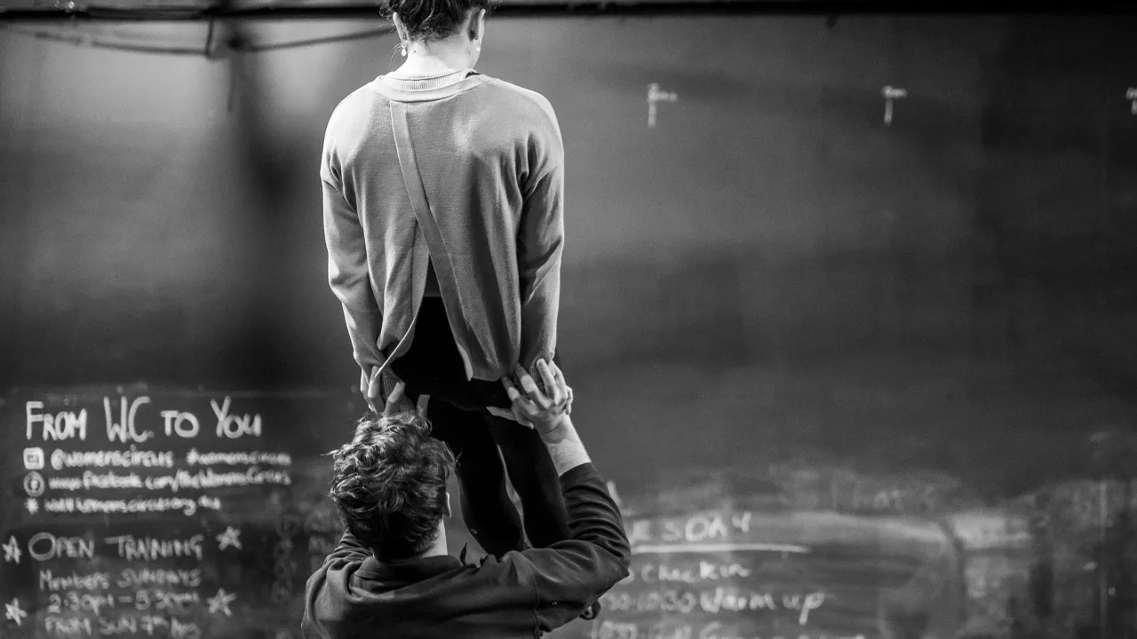 In a training space, two acrobats stand with their backs to the camera, facing a chalkboard covered in writing. One lifts the other as they practice a newly taught skill.