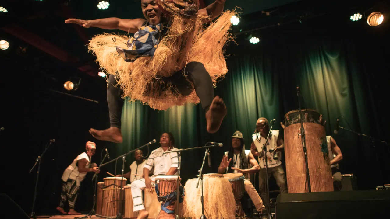 A band of African musicians play traditional drums, in the foreground, a dancer mid-air, arms & feet stretched out, wearing a grass skirt that is flared out in the jump.