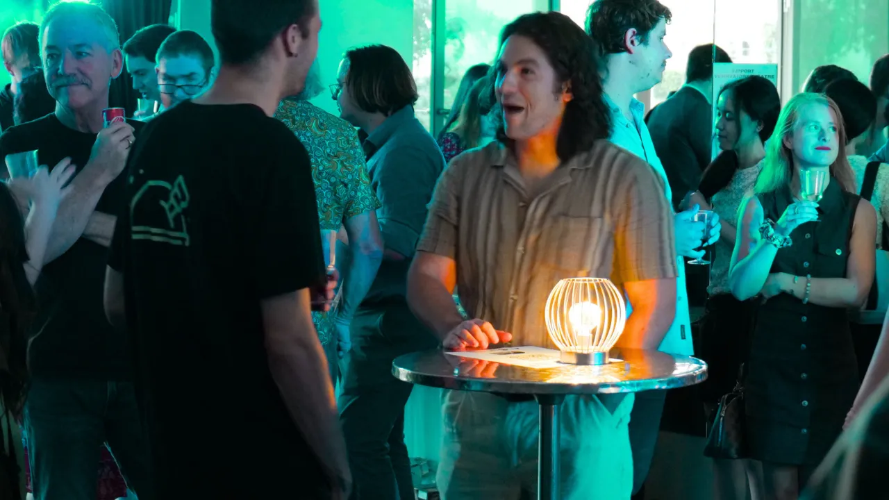 The image shows a group of people talking and laughing in a room with green coloured lighting. There is a dry bar table with a candle light on it that glows brightly among the green light.