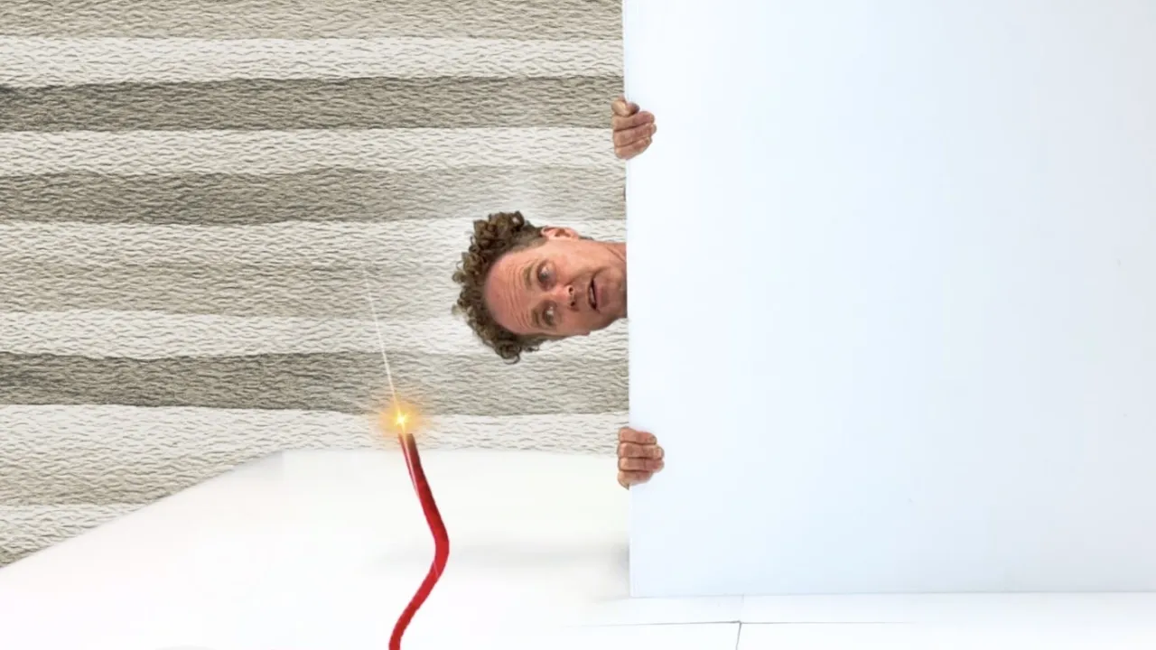 A man with curly hair is peeking around the edge of a large white rectangular piece of corflute, visible from about the eyes up. He is positioned behind this white corflute. In the foreground, there is a lit red fuse, with a bright flame at the tip. The fuse curves and coils in several loops. The image has been rotated 90 degrees, giving an unusual perspective where the man looks to be positioned horizontally rather than vertically.