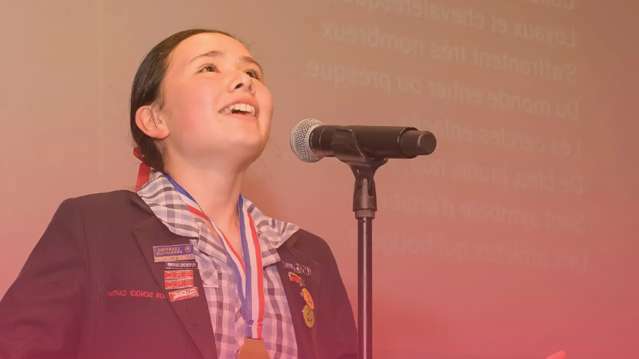 A student wearing a school uniform and blazer stands at a microphone, smiling and gesturing with open hands while reciting a poem on stage. She wears a gold medal around her neck, with a projected French poem visible in the background.