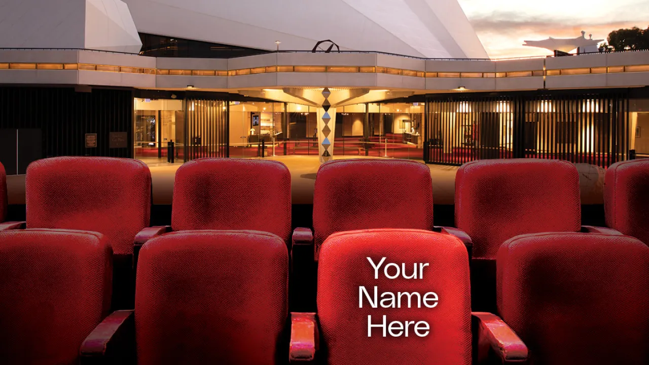Rows of plush red cinema seats are positioned in front of the entrance of the Adelaide Festival Centre, softly lit at dusk. One seat in the foreground stands out with white text reading “Your Name Here,” suggesting a sponsorship or donation opportunity. The scene conveys a warm, inviting atmosphere with the theatre’s glass doors glowing in the background.