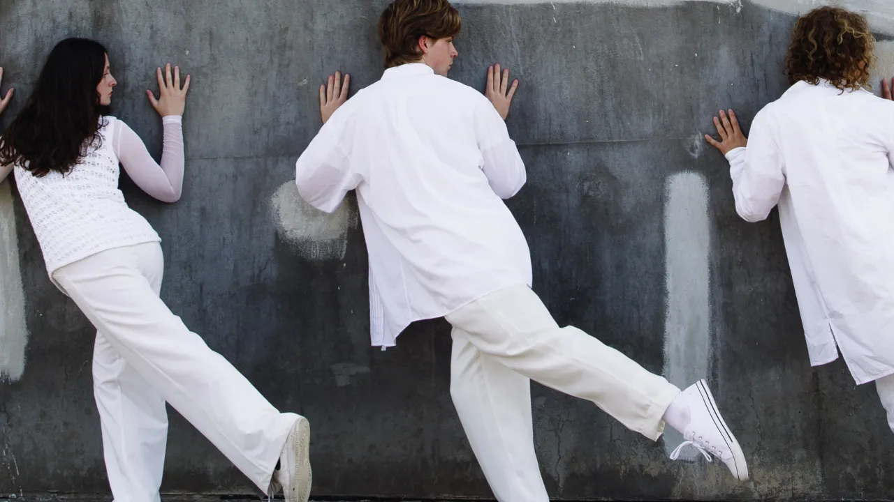 Three performers dressed in white clothing pose against a grey concrete wall, each pressing their hands to the wall and lifting one leg behind them. Their coordinated movements and minimalist outfits create a sense of balance and motion in a contemporary dance or performance art setting.