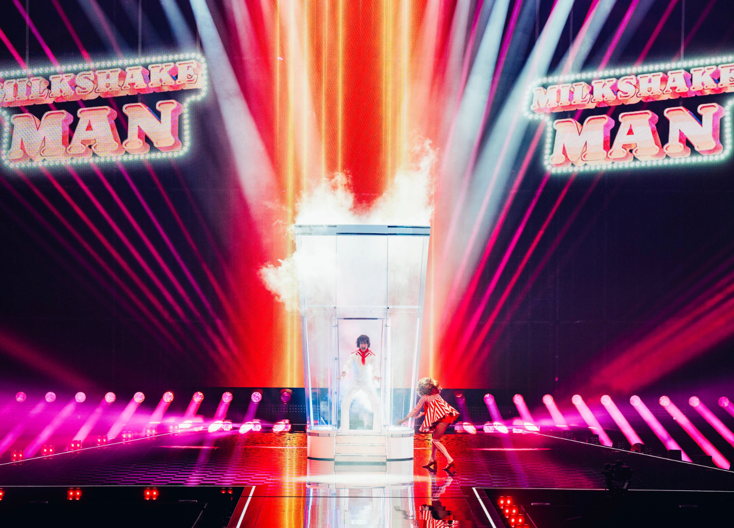 Go-Jo on stage at Eurovision, wearing a white jumpsuit and standing inside a giant blender. Projected on the colourful screen behind him is the words 'Milkshake Man'