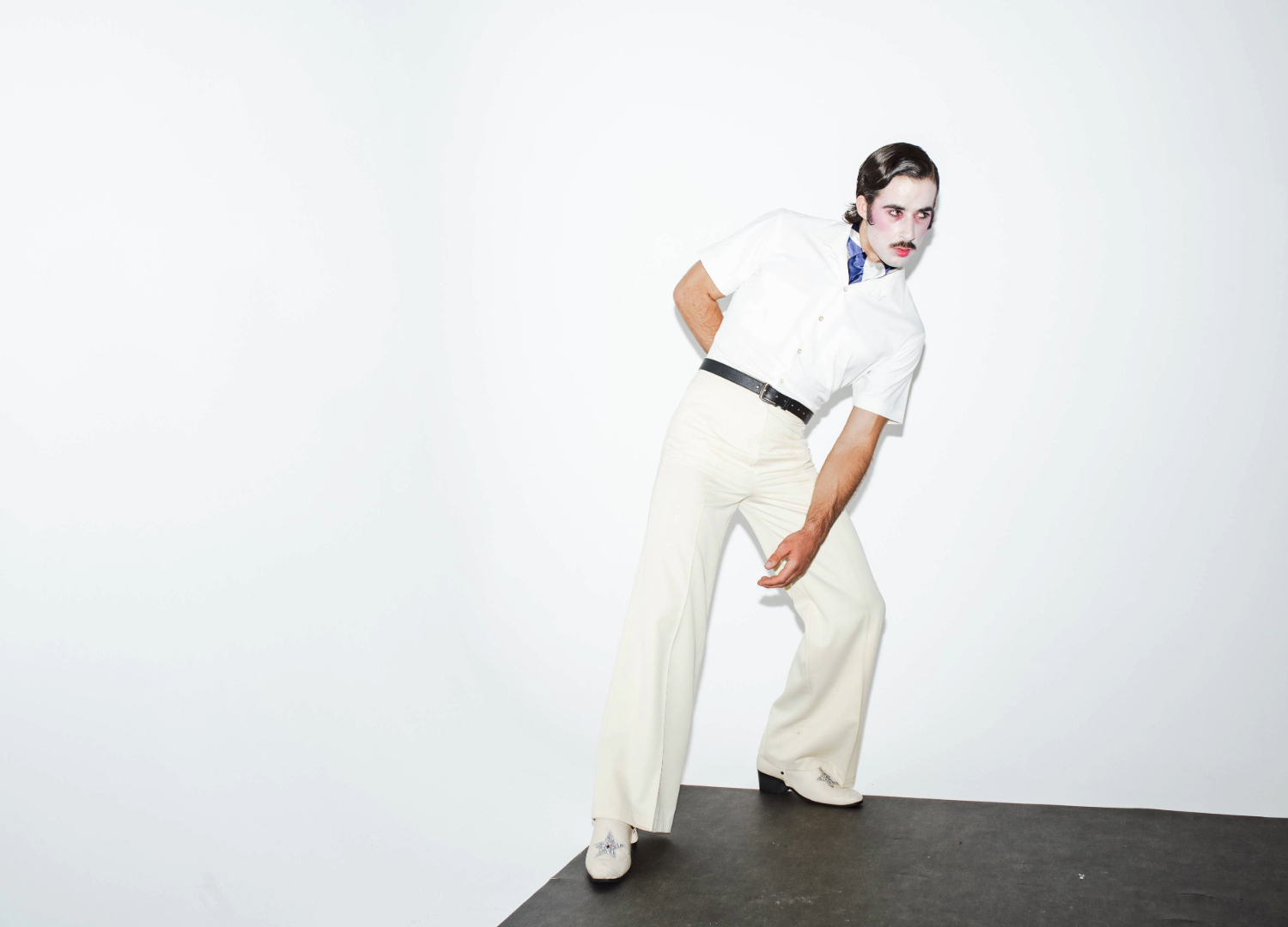 Go-Jo wears white slacks, a white short sleeve t-shirt and stands in the corner of a white room. His hair is slicked back, and his face is painted white with rosy cheeks and heavy eye makeup.