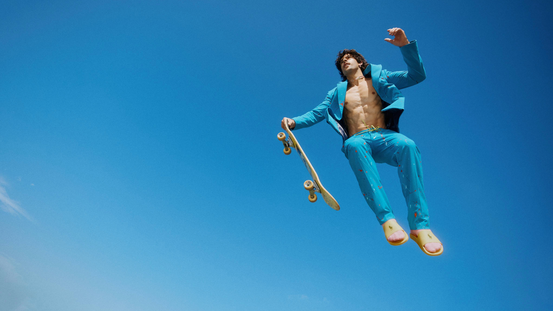 Go-Jo is wearing a blue suit, over his bare chest and is leaping in the air holding a skateboard. The vast blue sky behind makes it feel as if he is weightless in space.
