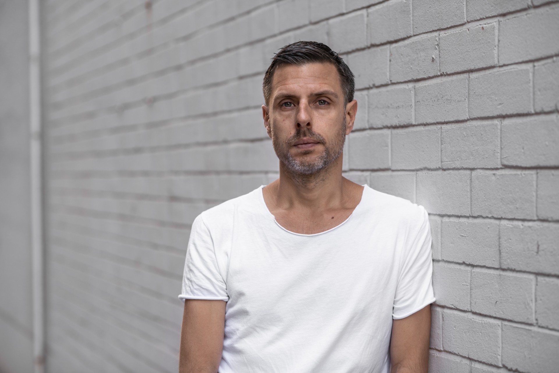 A headshot of Jonathan Jones. He is wearing a white t-shirt and leaning against a while brick wall.