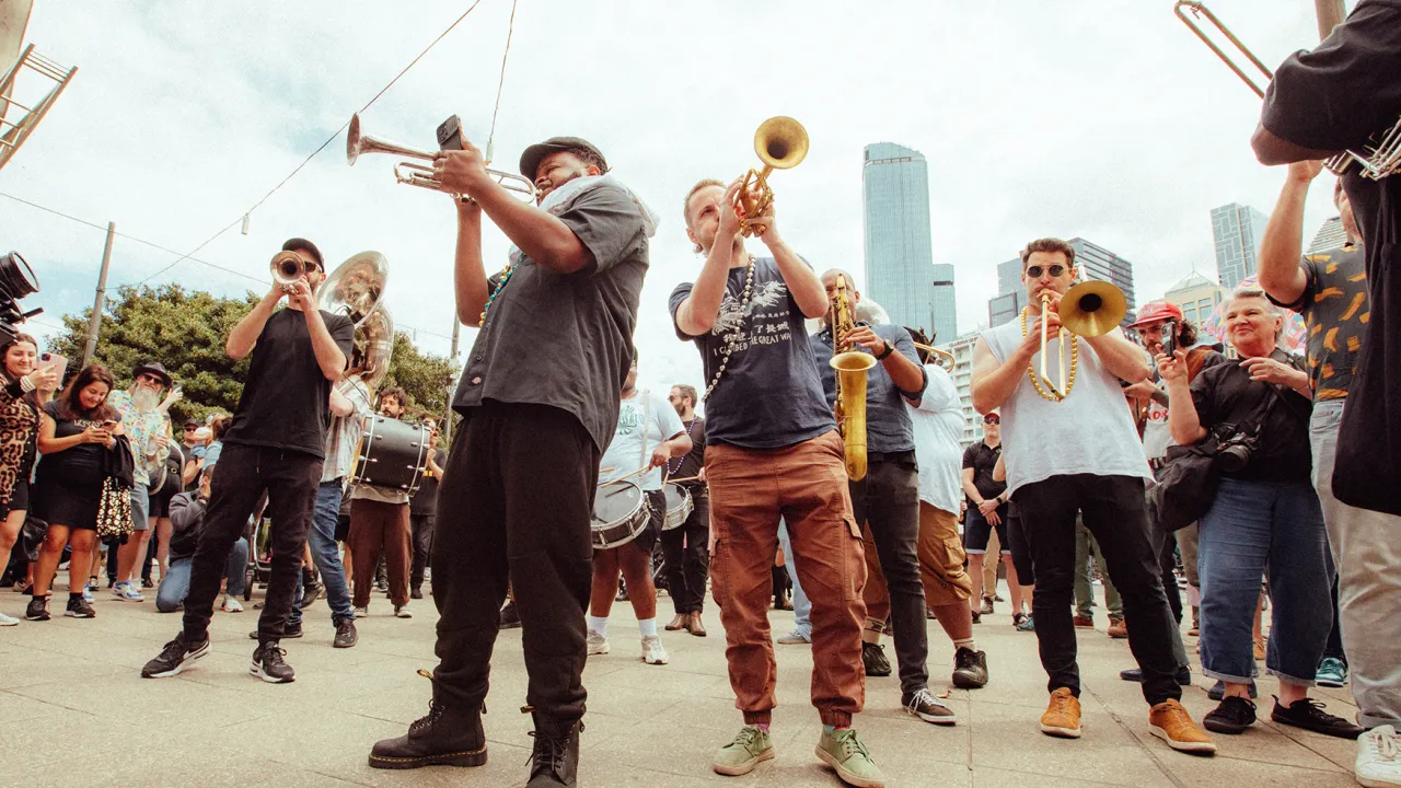 A jazz band stands in the street surrounded by a crowd of people. The camera is looking up at the performers.