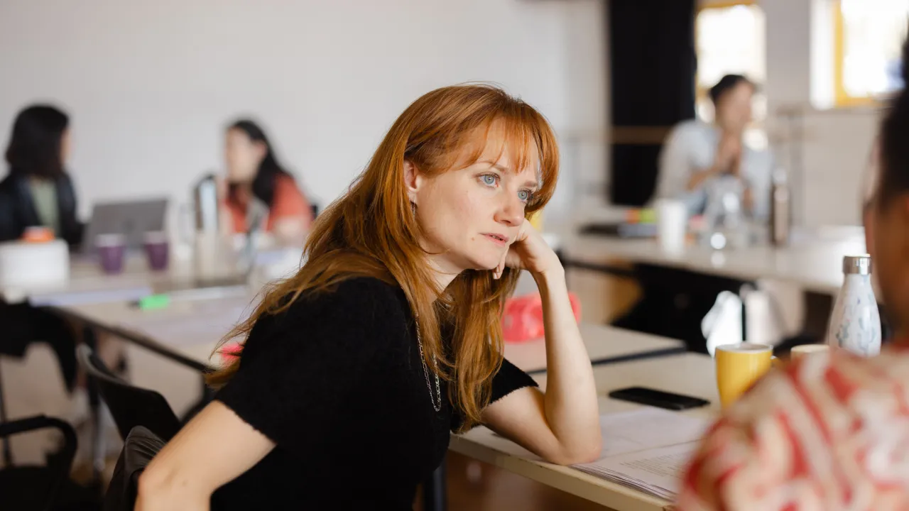 A woman with long red hair and a black top rests her head on her hand while sitting at a table in a workshop or meeting. Papers, cups, and water bottles are spread across the table. Other people are blurred in the background, engaged in conversation.