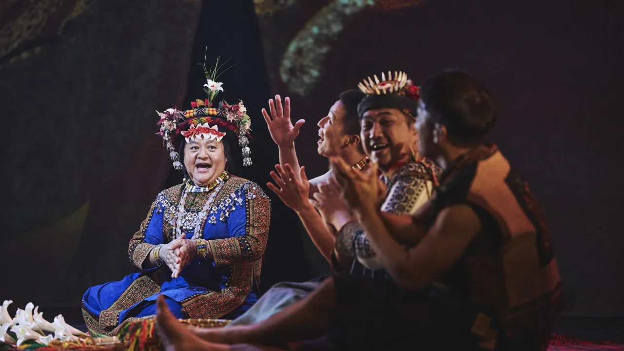 A group of performers in vibrant traditional attire sit on stage, engaged in song or storytelling. One performer, dressed in a bright blue embroidered outfit with an elaborate floral and beaded headdress, claps hands while others beside them smile, gesture, and sing enthusiastically. The scene is lively and expressive, with cultural costumes and joyful interaction highlighted against a dark backdrop.