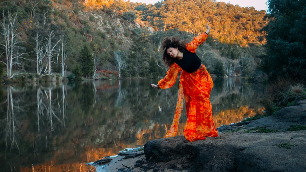 A female First Nations dancer with curly hair moves in a vibrant orange top and skirt in front of a river and australian bushland.
