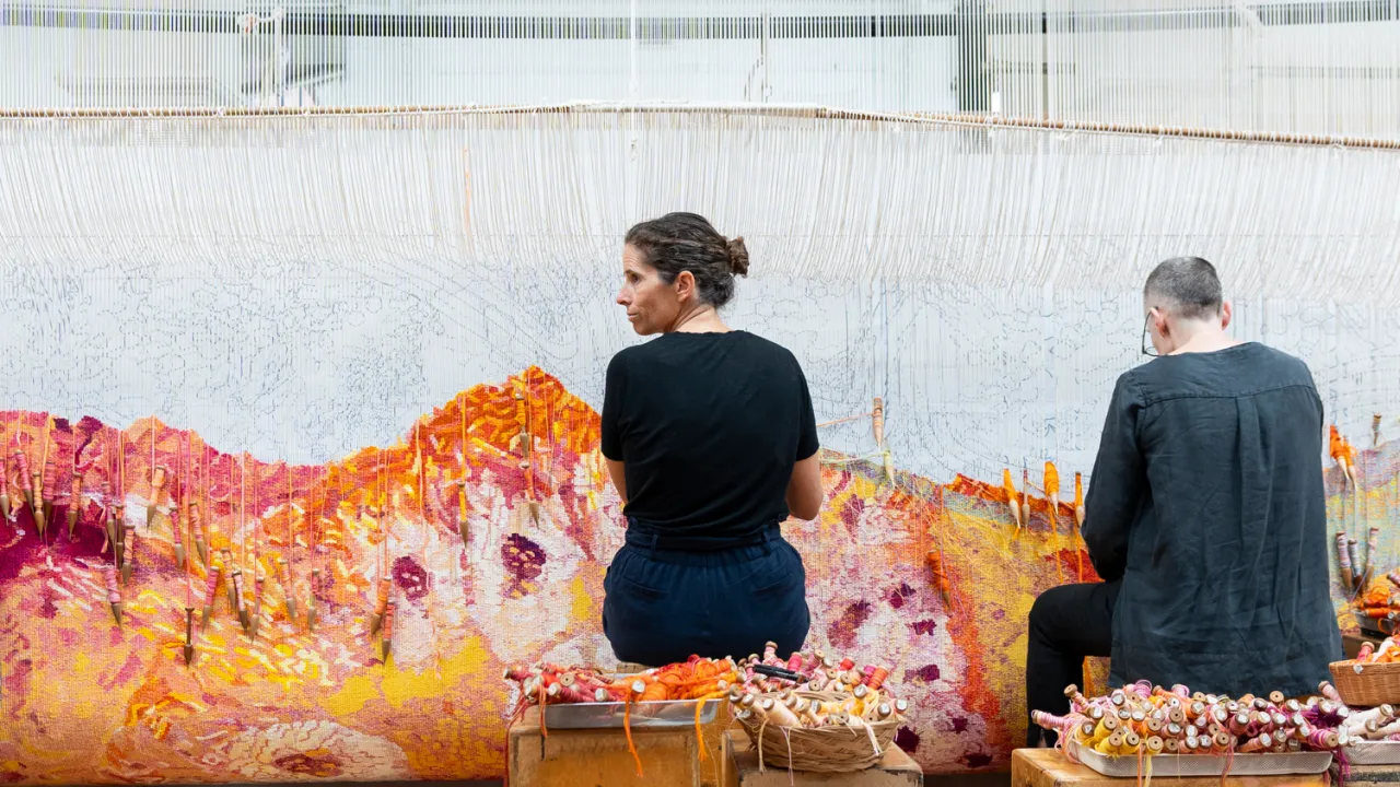 Two weavers sit side by side at a large loom, weaving a tapestry. They are surrounded by baskets of colourful yarn.