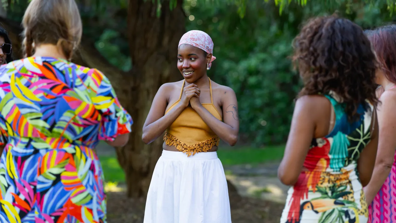 A young woman in a mustard halter top and white skirt smiles warmly with hands clasped, standing outdoors in conversation with three people in colourful clothing.