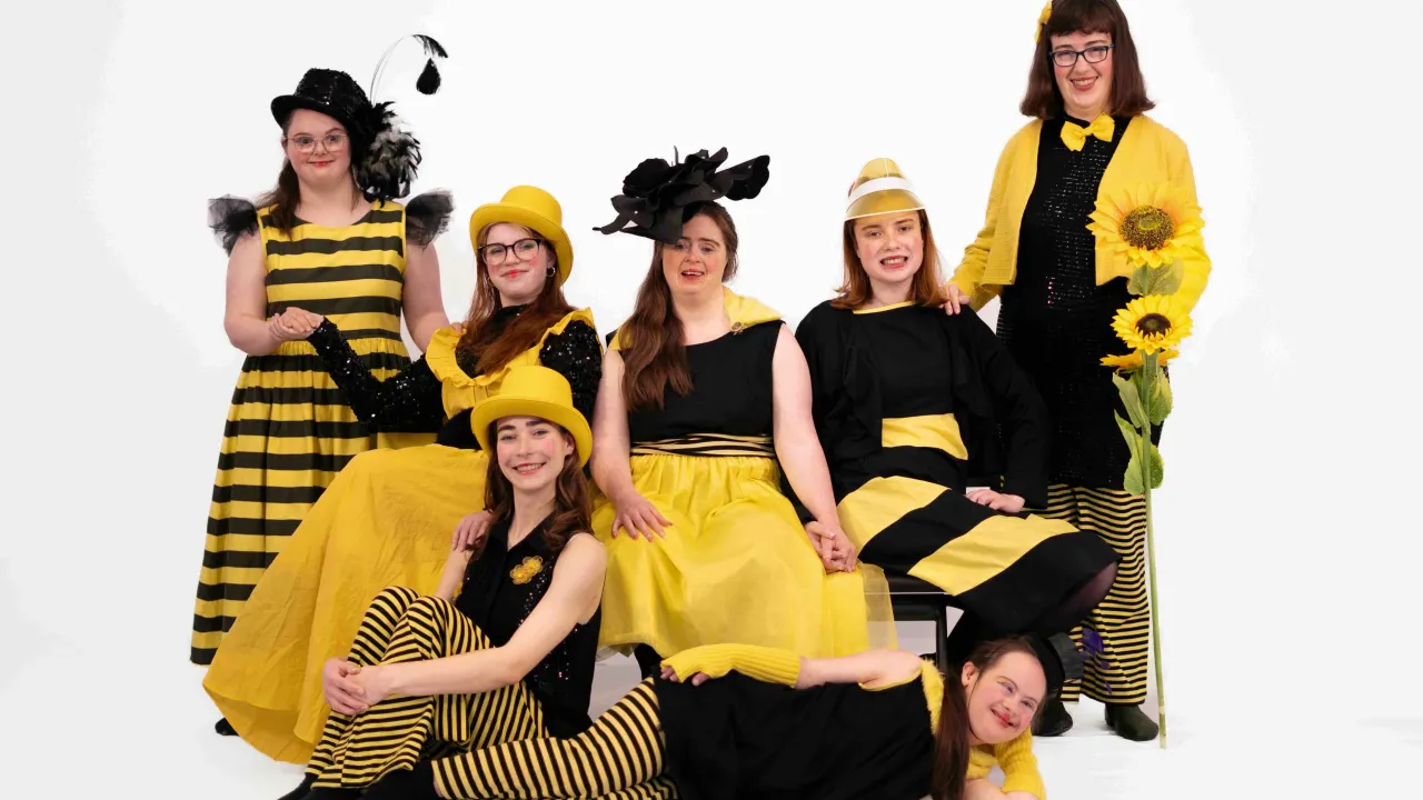 8 women dressed in black and yellow costumes and hats sitting and standing in a white space looking at the camera and smiling.