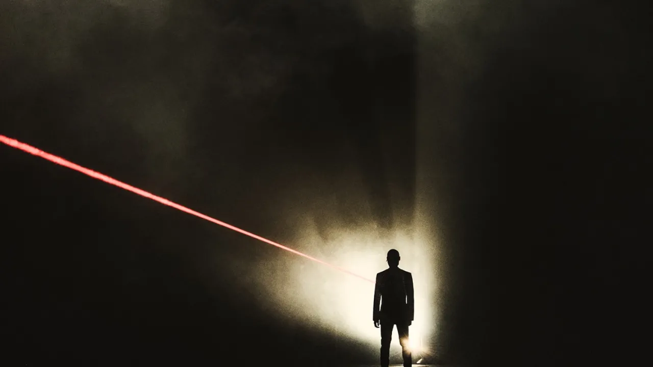 An image of a male person standing in the dark with a bright red laser beam pointing at them. The image is a work by Matthew Sleeth called A Drone Opera.