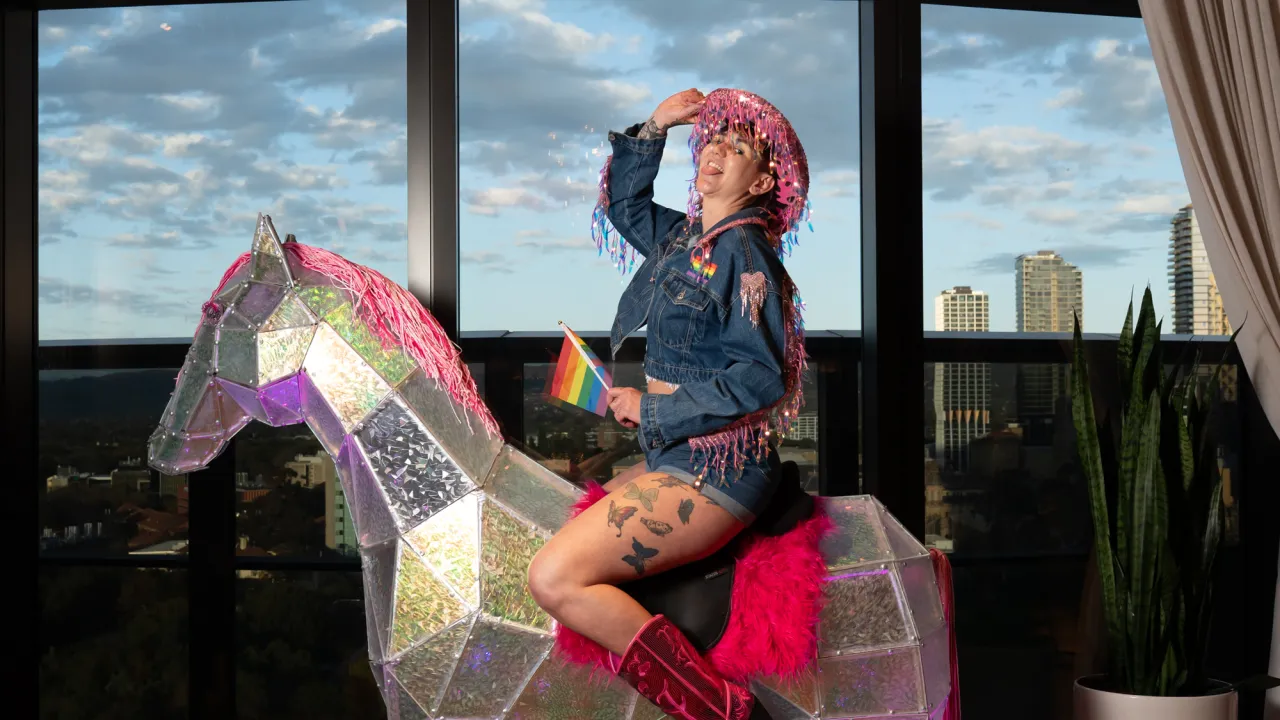 A person in a sparkly pink cowboy hat, denim jacket with tassels, and pink boots poses on a glittery geometric horse with pink accents, holding a rainbow pride flag and smiling in front of a city skyline.