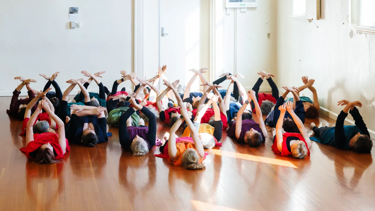 Older dancers lay on their backs, their arms cross above their bodies, like birds flying in the sky.