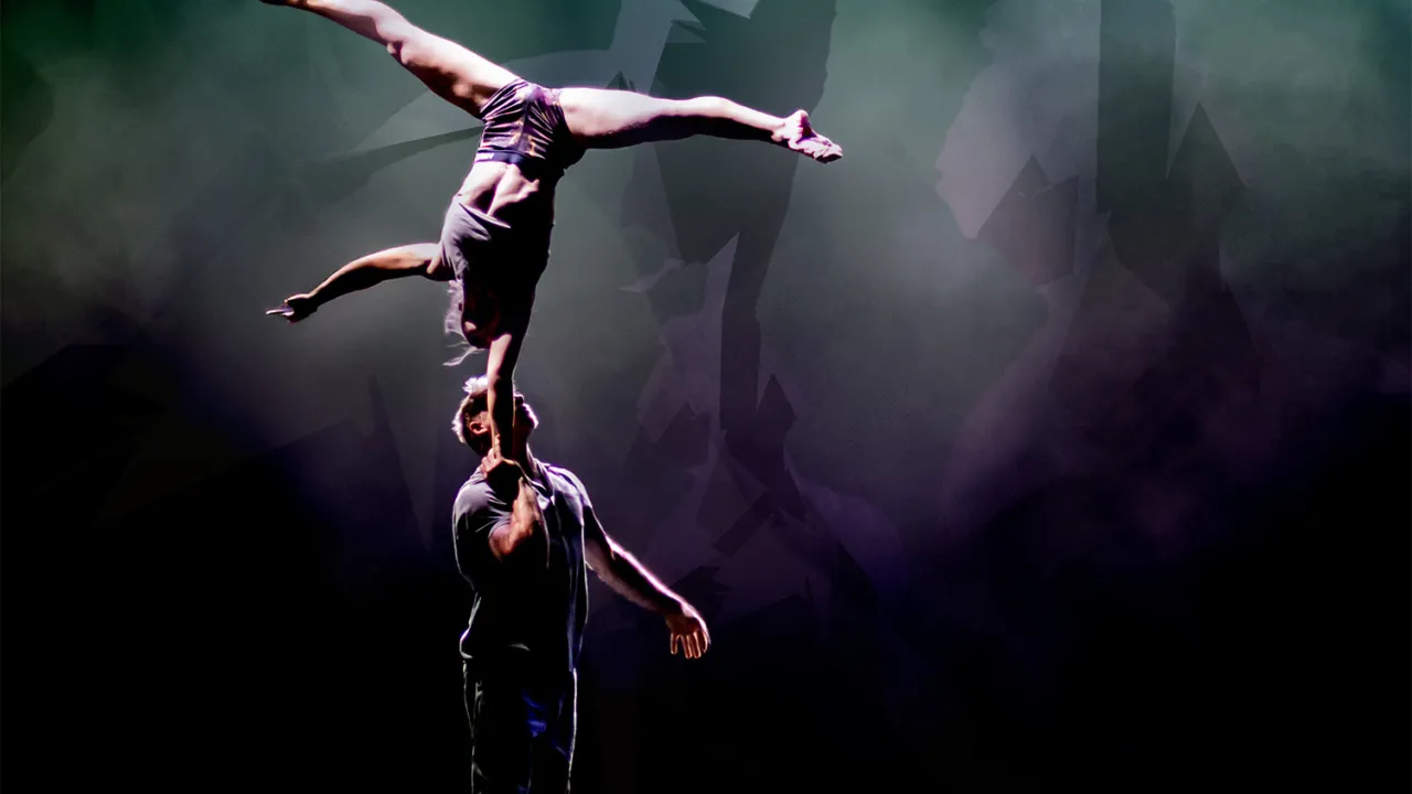 Two acrobats in grey clothing with a shadowed background of dark greens and purples. One acrobat is standing on the ground holding the other in a one arm handstand.