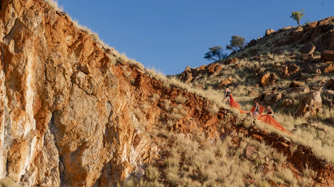 3 women wearing black swim wear walk up a small ridge in the desert. They are carrying a piece of bright orange plastic and a blue rope, the rock face is burnt orange and white, and the sky is bright blue.