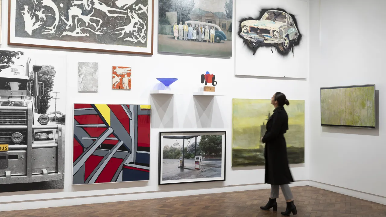 Visitor wearing in black boots, grey jeans and a black jacket is standing in front of a salon hang of artworks including paintings, photographs, small sculptures on shelves and a moving image work (still image) on a monitor.