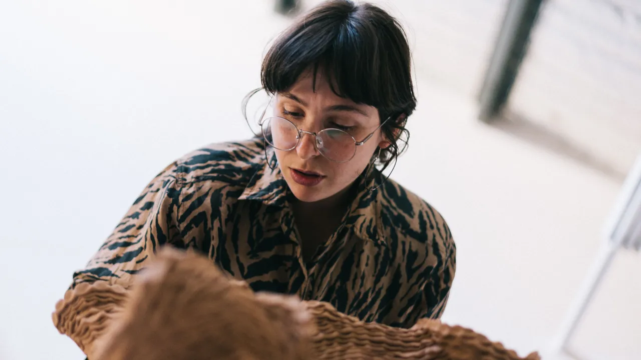 Artist working on a ceramic work. They are seen leaning over the work. The person has brunette hair, glasses, and an animal print shirt. The artwork is tan coloured ceramic in the foreground. The background is minimal studio environment