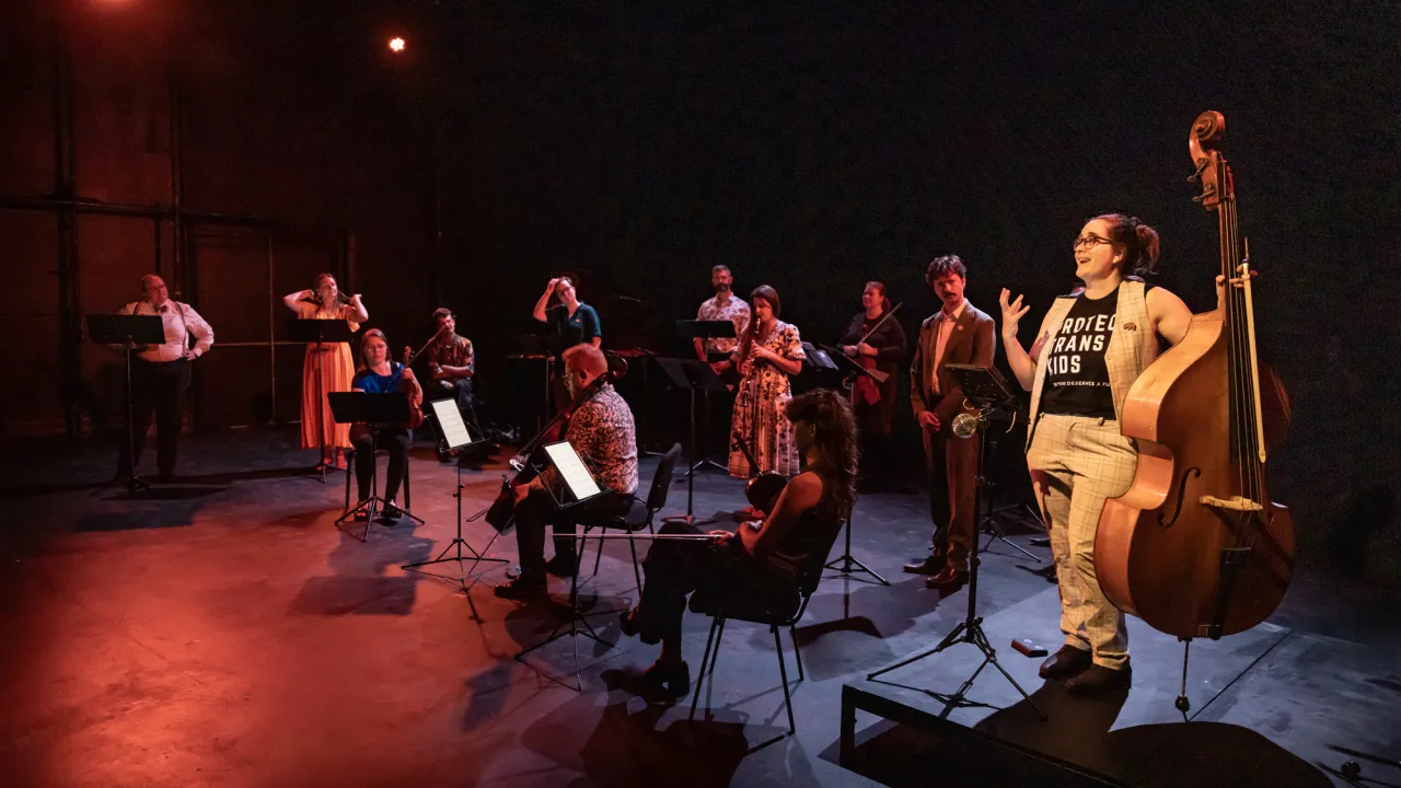 A group of people are scattered across a darkened stage. Some have instruments. All are dressed colourfully. A woman stands in the light at the front holding a double bass and talking animatedly wearing a tshirt that says "protect trans kids"