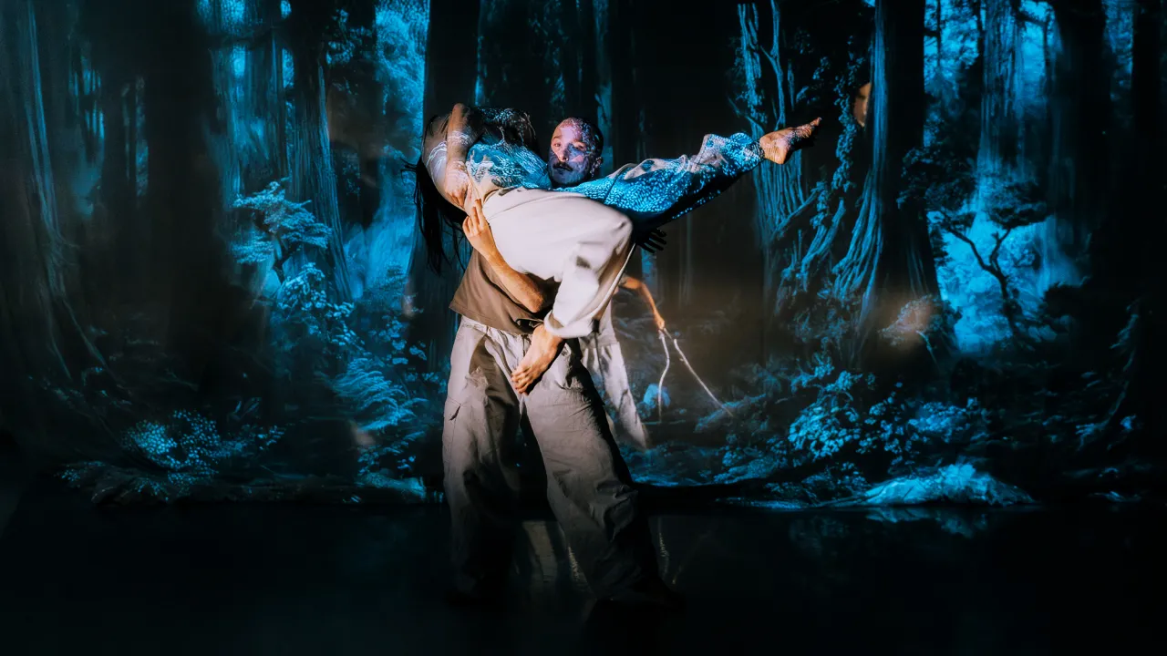 Two dancers performing, one lifted in the air by the other, surrounded by projected forest imagery, creating a dramatic and immersive scene