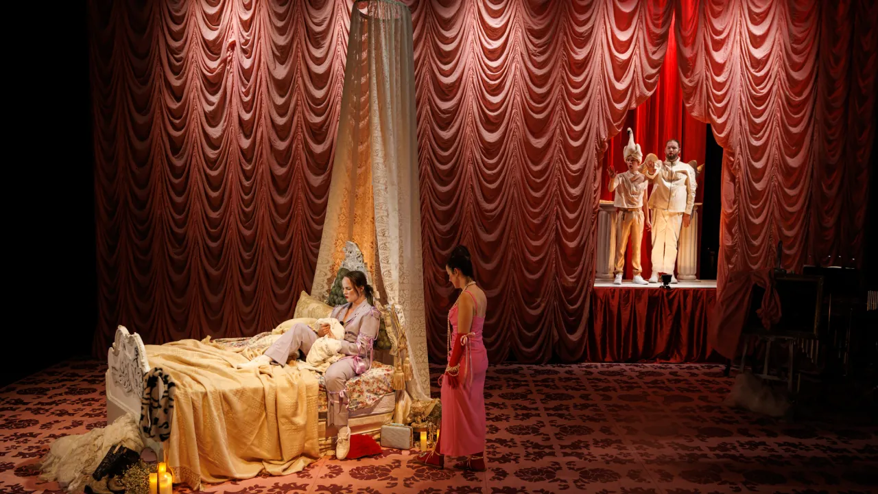 Stage screenshot of 2022 Iphis Production. Opera by Elena Kats-Chernin and directed by Katy Maudlin. Image features a wide stage screen shot with a pink curtain background. A mother is arguing with her daughter, who is sitting on an elaborate bed. There are two god-like figures in the background of the photo.