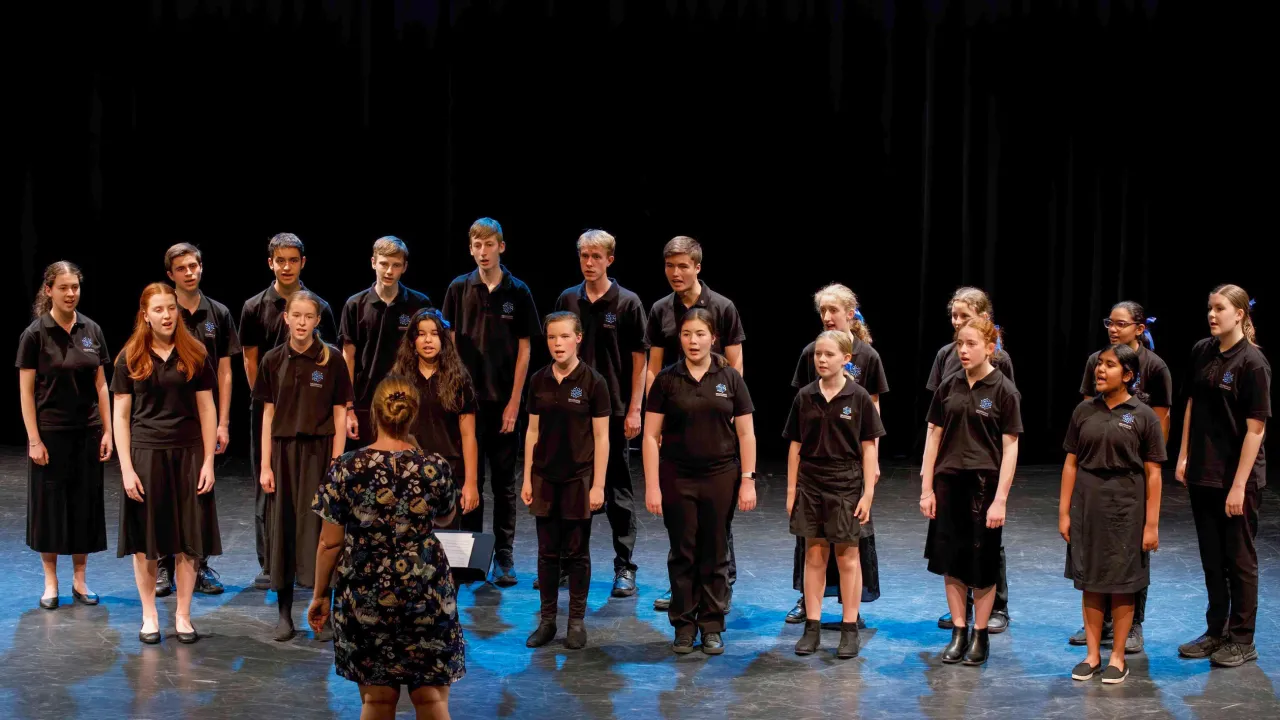 The image features 19 young people standing in two rows, showing their faces. There is also a backview of the conductor standing in front of them. The choristers are dressed in their performance uniform, black polo tops with MYC's logo and black bottoms. There is a blue background. They are singing.