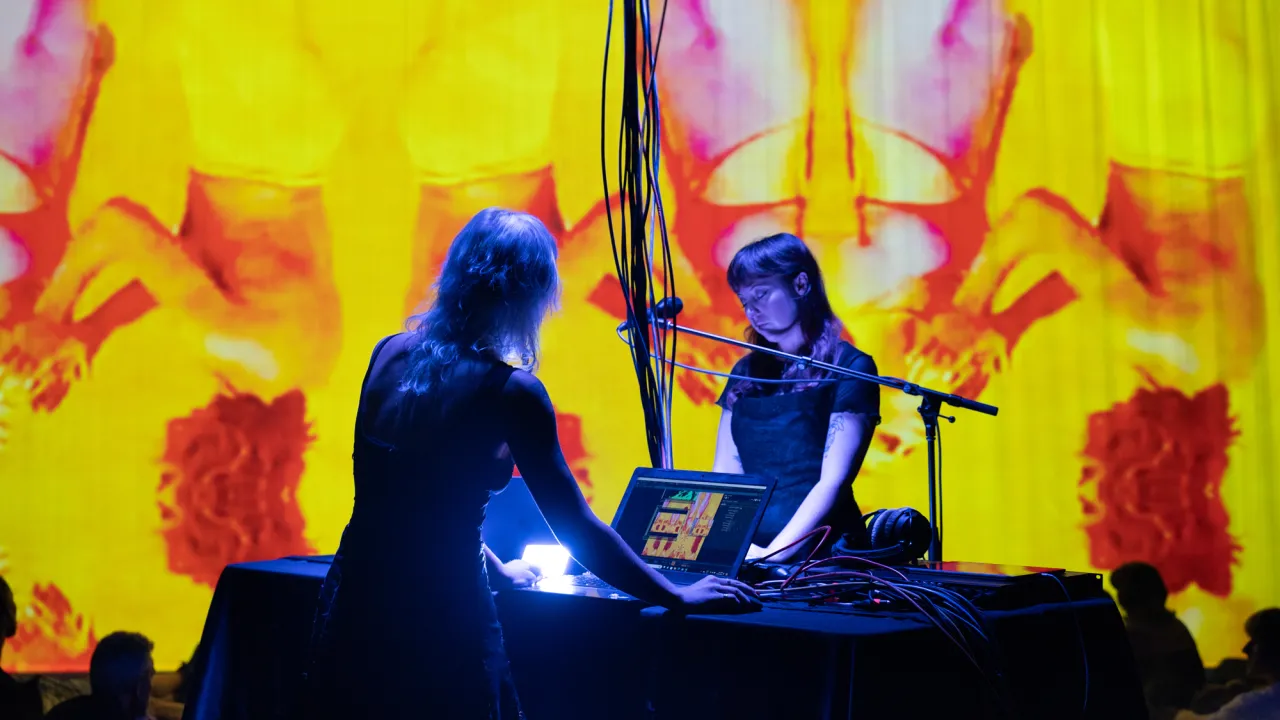 Two artists interacting with audio equipment and computers in front of an extremely large and vibrant yellow, red and fuchsia digital art piece
