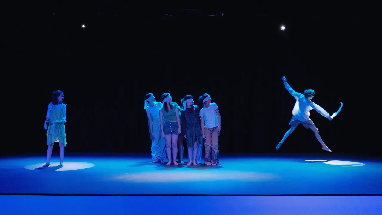 A theatrical performance scene is lit with dramatic blue lighting. 12 performers are on stage. On the left, a young girl in a light green costume stands alone under a spotlight, holding a piece of fabric. In the center, a group of 10 individuals stands closely together, all blindfolded and barefoot, facing slightly to the right. On the far right, a male dancer is captured mid-leap, with one arm raised and a strip of fabric flowing in the air. The background is entirely black, highlighting the performers und