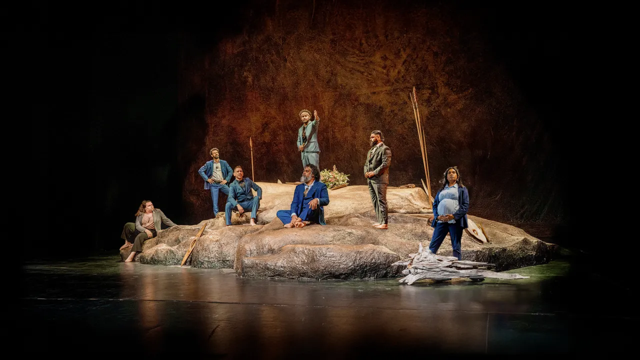 The 2025 Cast of The Visitors by Jane Harrison. 7 clan leaders stand on a rocky escarpment. Watching some strangers on a boat sail into Sydney Harbour