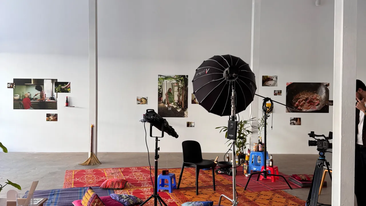 A spacious, white-walled, high ceiling gallery features a photographic exhibition showcasing cooking methods and food traditions from Asian Australian families. Large and small photographs are arranged across the walls, depicting scenes of people preparing meals and close-ups of dishes being cooked. In the center of the gallery, a separate video production setup is arranged for a campaign interview series. This includes lighting equipment, cameras on tripods, and a black chair placed on top of layered patte