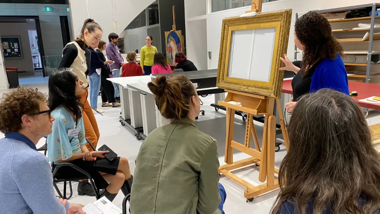 The image shows a group of people gathered in an art conservation lab. They are participating in a workshop focused on caring for art works. In the foreground, several seated attendees are attentively watching a woman in a blue cardigan, who is standing next to an easel holding an empty, ornate gold picture frame. In the background, other people are engaged in discussion of a painting.
