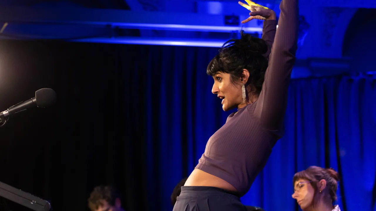 "Comedian Vidya Rajan performs during Poetry Month 2025. She is on stage behind a microcphone. She has dark hair in a bun, sparkly dangling earrings, and a purple cropped top. She reaches upward expressively. Other performers are seated behind her under blue stage lighting. "