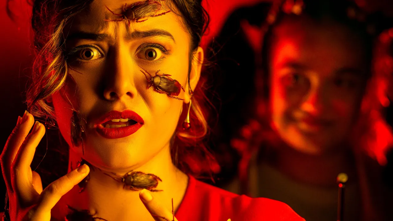 A face is seen in shock, with their fringe curled falls to the side of their eyes and cheek, they also have their hand hand towards their face. Their face and hands are covered in cockroaches while drenched in red and yellow light. There is a blurry shadow of another face behind them somewhat grinning while holding a remote control device with a large antenna.