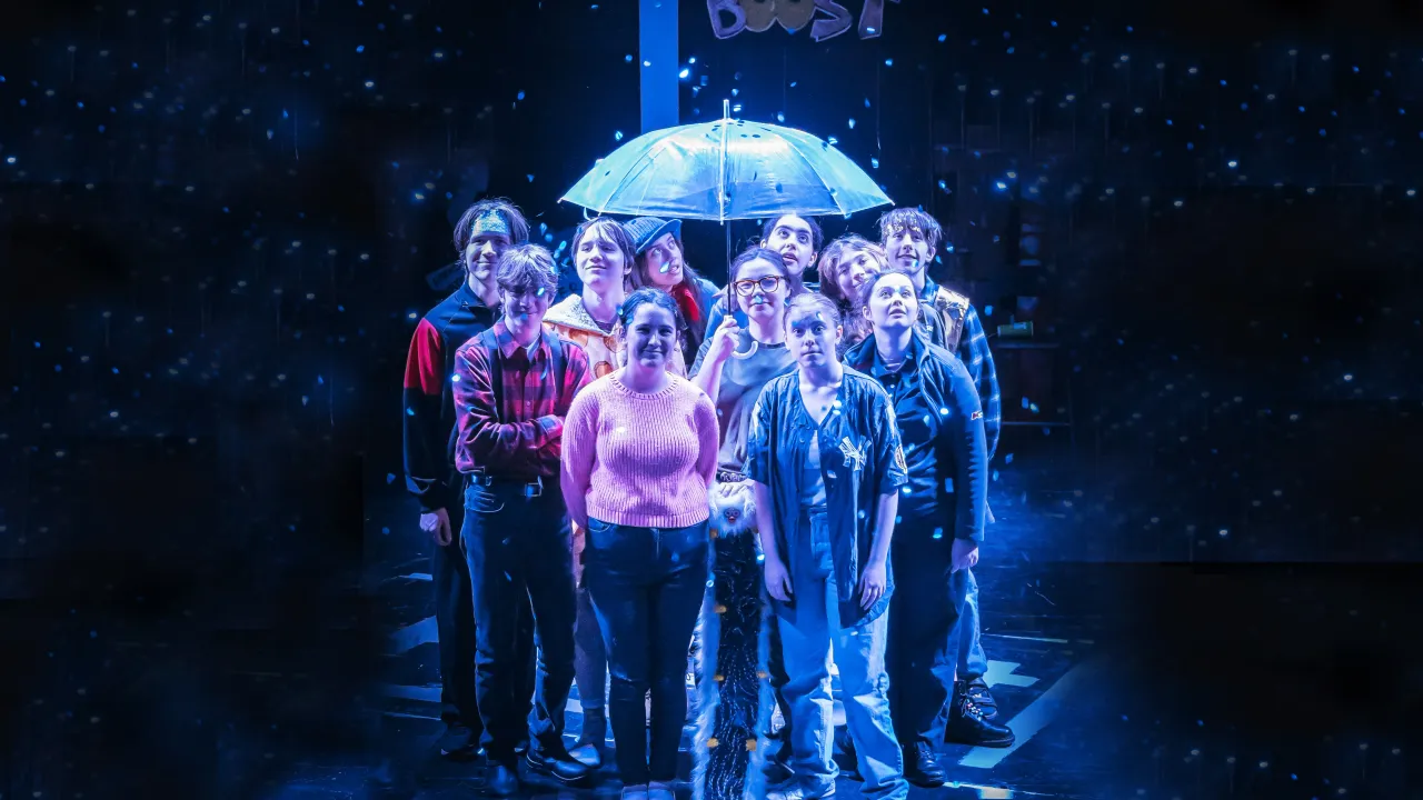 Shopfront's Senior Ensemble on stage in When I Grow Up. They are under an umbrella as confetti rains.