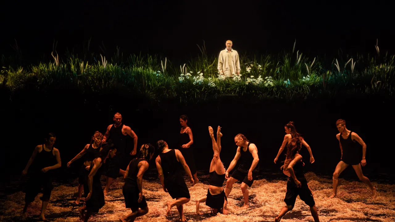 Twelve dancers, dressed in black, perform on a stage covered in hay. The central dancer is upside-down in a headstand and behind the dancers a male singer stands on a raised platform surrounded by plants and flowers.