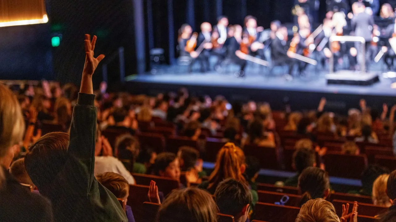 A child raises their hand enthusiastically to ask a question of musicians on stage after a performance.