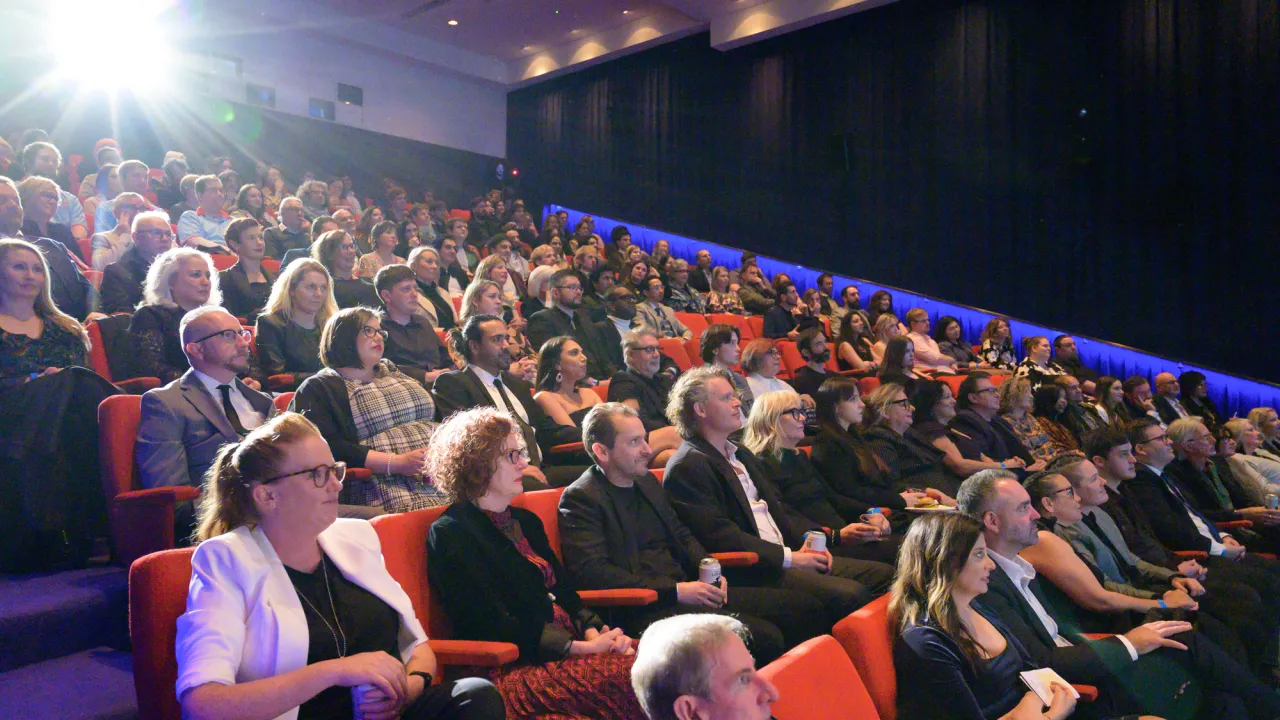 A full audience sits in a cinema with the strong light from the projector shining from the back of the theatre