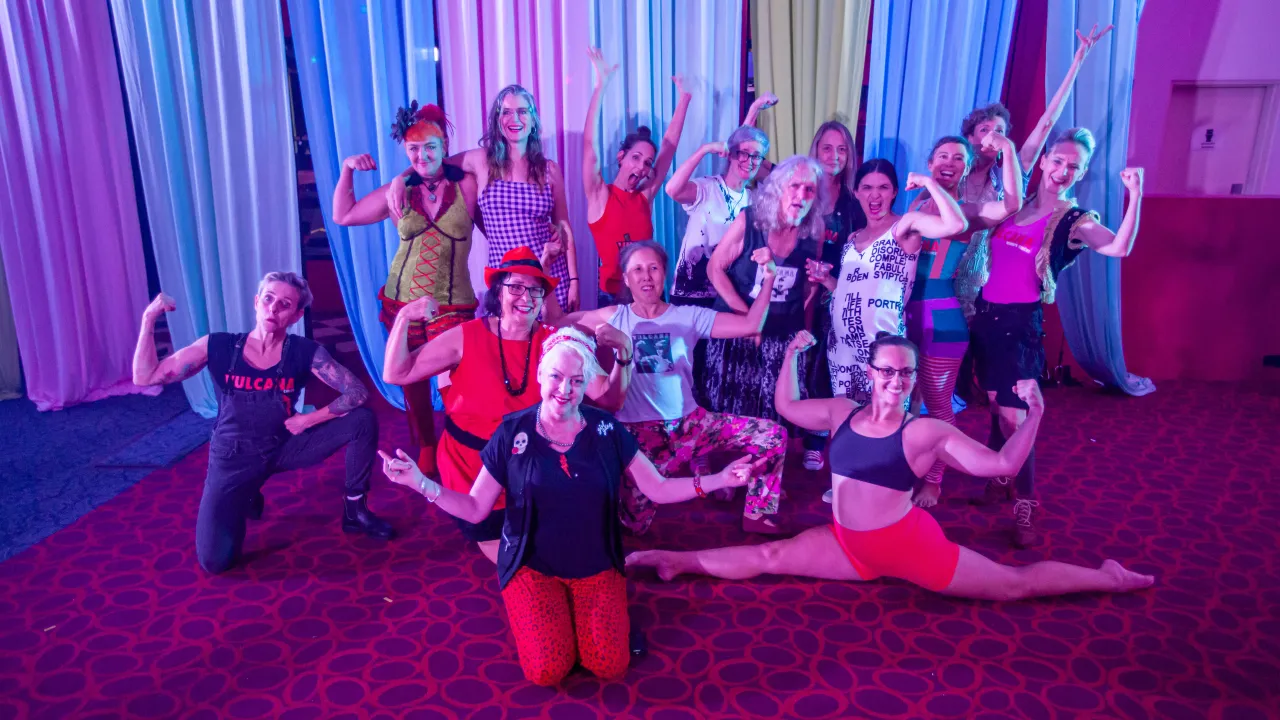 A photo of 15-20 people in feminine and gender-ambiguous clothing posing in a celebratory way in front of a curtain with red lighting. Some are crouched, some are flexing their arms to show their muscles, some are standing and doing the splits. All look happy and proud.
