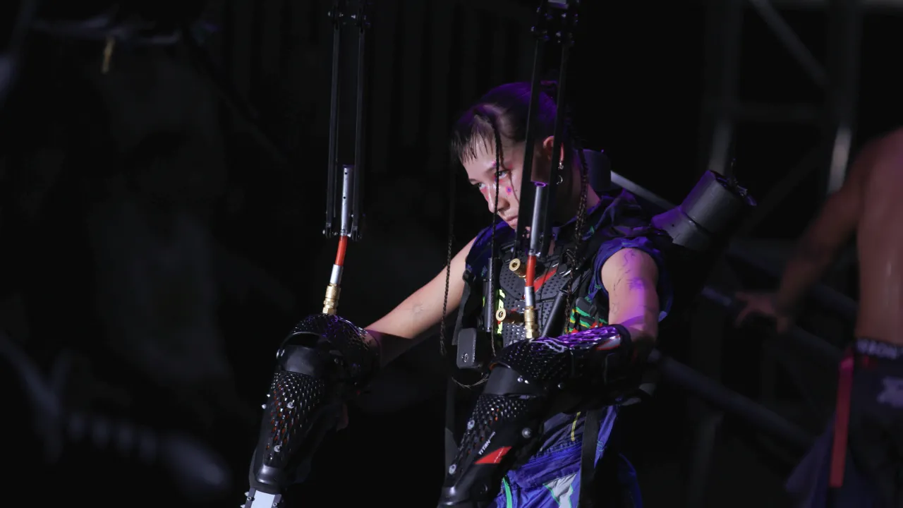 A performer stands under dramatic lighting wearing a futuristic exoskeleton-like costume with mechanical arm extensions. Their intense expression and forward-leaning posture suggest focus and strength. The scene is dark with metallic structures and another person partially visible in the background, creating a moody, high-tech atmosphere.