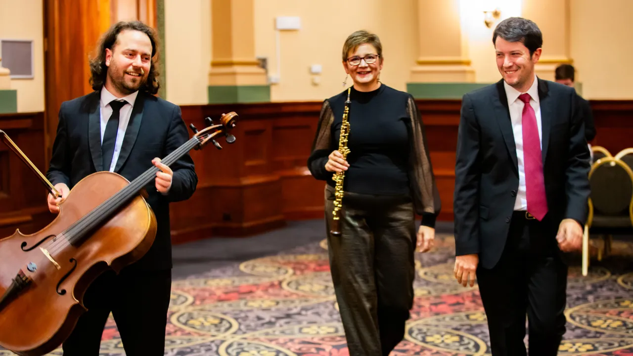 Three musicians walking together indoors. The man on the left is holding a cello and bow, the woman in the centre is holding an oboe, and the man on the right is smiling. All are dressed formally in dark attire, with the two men wearing suits and ties. They are walking across a patterned carpet in a warmly lit room with wood paneling and chairs in the background.