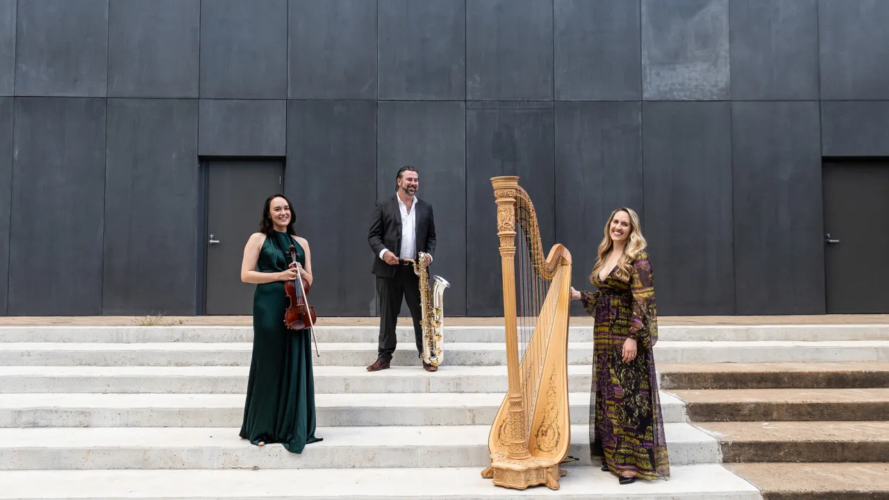 Three musicians are featured in a black background wall, two females with string instruments and a mae with a brass made instrument.