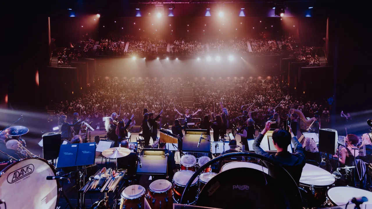 A full orchestra performs on stage in a large concert hall, viewed from behind the percussion section toward the audience. Musicians play drums, brass, strings, and woodwinds under dramatic stage lighting, with beams of purple and gold light illuminating the space. The conductor stands at the centre with arms raised, while a packed audience fills the tiered seating, creating an atmosphere of excitement and grandeur.