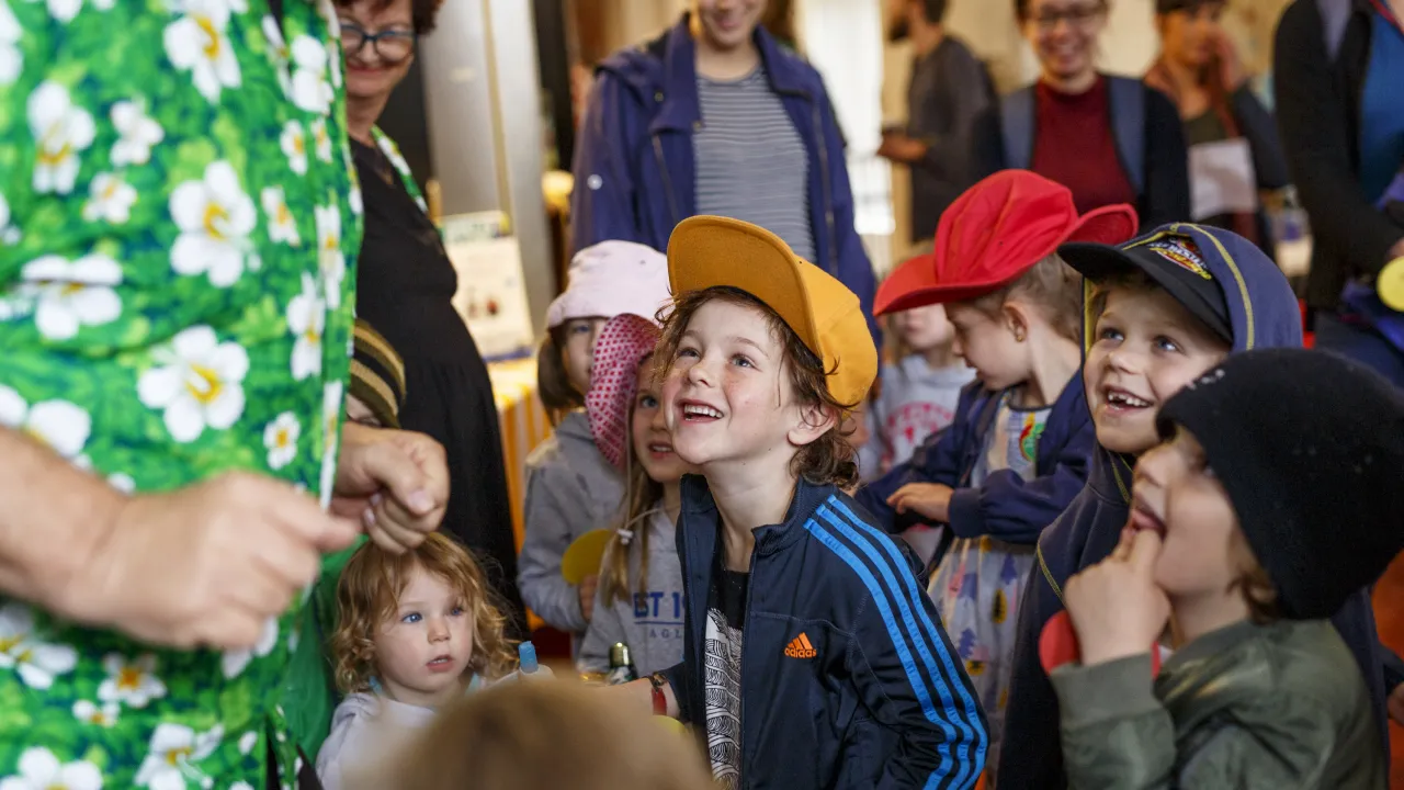 A group of young children, many wearing colourful hats, look up with wide smiles and excitement toward an adult in a bright green floral shirt. The scene takes place indoors, with several adults in the background watching and smiling. The atmosphere feels lively and joyful, as if the children are watching a performer or storyteller.